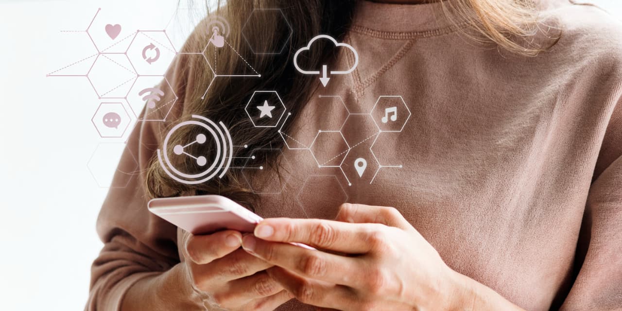 A person holding a pink smartphone with both hands, interacting with the screen. Over the image, digital icons in white tones are superimposed, representing technological and connectivity elements, such as cloud, sharing, chat, location, star, music, Wi-Fi and heart. The person is wearing a light pink sweatshirt, and the background is neutral, clear and blurred, highlighting both the cell phone and the digital icons. The image conveys the idea of ​​connection, technology, digital transformation and the use of mobile devices for browsing and interacting online.