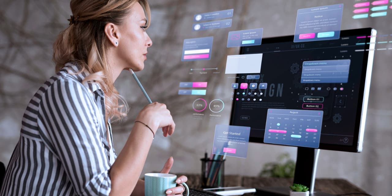 The image shows a woman sitting in front of a computer, focused on her work. She holds a pen close to her face and a mug in her other hand, while looking at the screen filled with holographic digital interface elements, such as menus, buttons and graphics. The scene suggests a modern work environment, with a focus on technology, user interface design (UI/UX) and digital innovation.