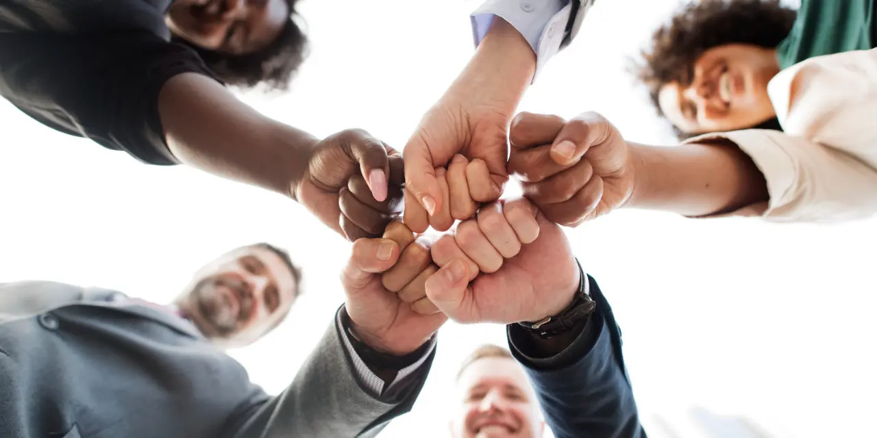The image shows a group of people in a circle, viewed from below, clasping their fists in the center in a gesture of unity and teamwork. The hands are of different skin tones, representing diversity. Everyone is smiling, conveying a positive atmosphere of cooperation and partnership. The people wear formal and casual clothing, suggesting an inclusive and friendly professional environment. The background is light, with a white sky in the background, highlighting the collective gesture.