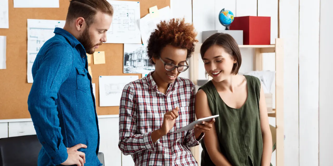 Three people in an office environment looking together at a tablet and smiling.