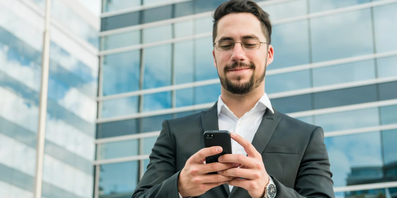 A young man with a beard and short hair is wearing a black suit with a white shirt and glasses. He smiles slightly as he looks at a smartphone he holds in both hands. In the background is a modern office building with a glass facade reflecting the blue sky and a few clouds. The scene conveys a corporate and professional atmosphere, with a focus on technology and connectivity.