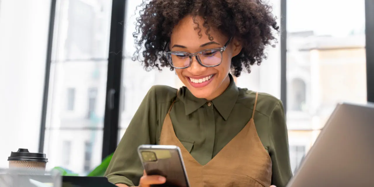 Mujer sonriente con gafas usando un smartphone en una oficina luminosa.