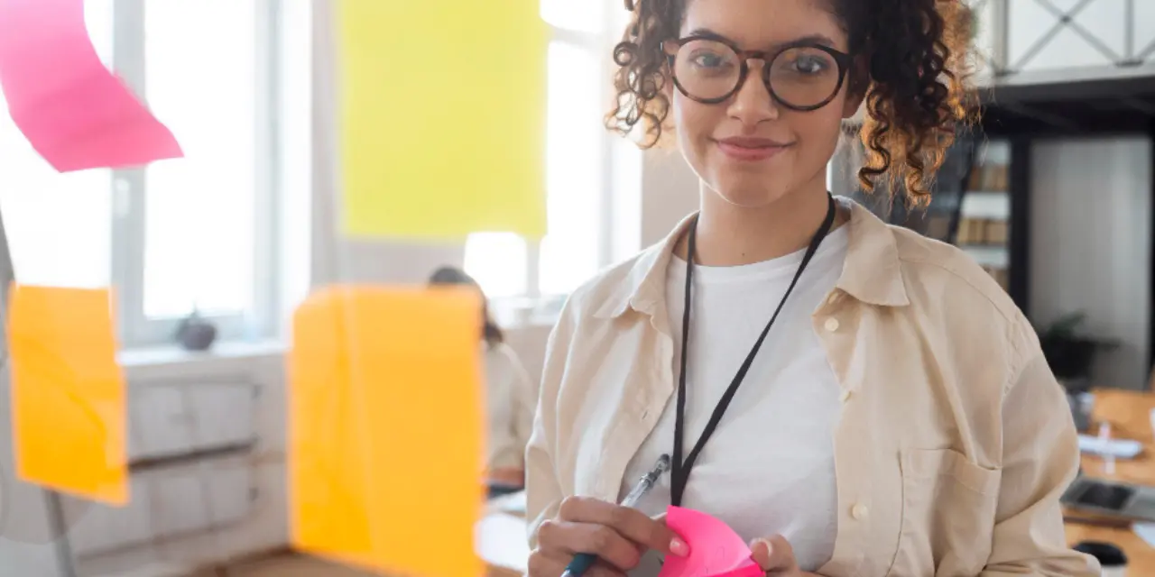 Pessoa sorrindo em escritório com post-its coloridos em um quadro de vidro.