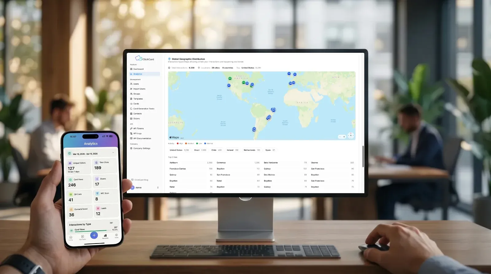 A first-person view looking down at a wooden desk in a busy, modern open-plan office. A hand in the foreground holds a smartphone displaying the ClickCard mobile app analytics dashboard for the period of March 16, 2026 to April 15, 2026. The mobile screen details key metrics such as "Unique visitors" (127), "Total clicks" (189), and "Card views" (246). In front of the hand, a large desktop monitor displays the full ClickCard web analytics dashboard, featuring its main "Global geographic distribution" map. The text on the monitor screen lists global interaction counts (e.g., Total interactions: 202) and a list of top cities. A wireless keyboard and mouse sit on the desk. Two colleagues are visible working at desks in the softly blurred background of the office.