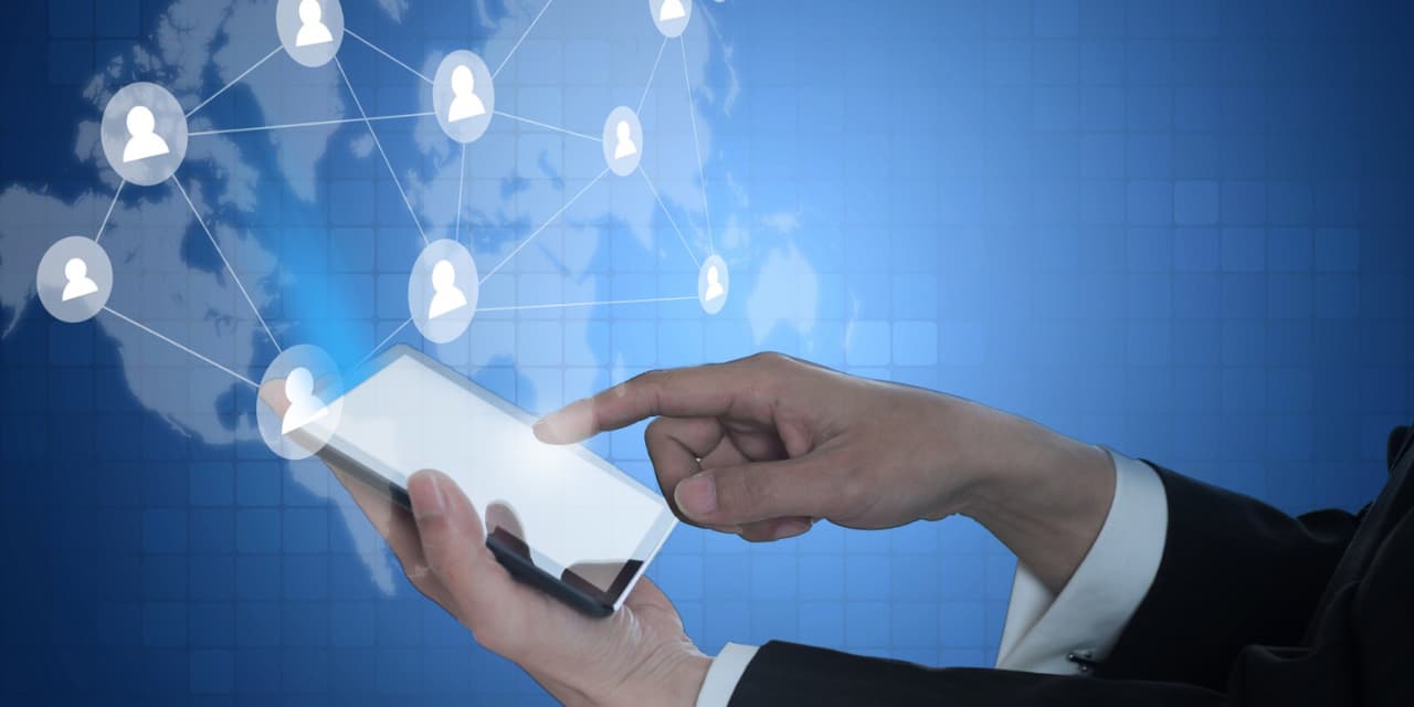 A person using a tablet on a technological blue background. On the screen of the device and around it appear icons of people in circles, connected to each other by white lines, representing a network of connections. In the background, there is a blue world map, suggesting the idea of global connection and digital networking. The person is wearing a black suit, indicating a professional or business context.