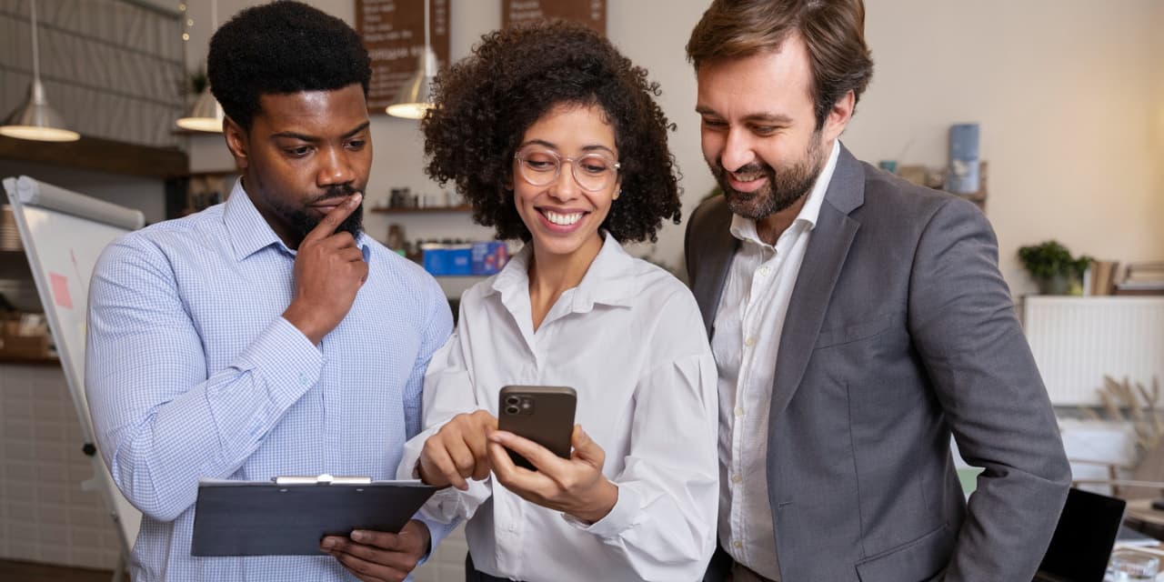 Three people are standing in a modern corporate setting, smiling and looking at a smartphone screen. In the center, a woman with curly hair wearing glasses and a white shirt is holding her phone and showing something to her colleagues. On the left, a man with a thoughtful expression is holding a clipboard and looking intently at his phone. On the right, another man in a gray suit and white shirt is smiling while also looking at the screen. In the background, you can see part of an office or coffee shop, with cozy decor, pendant lights, and a menu board. The scene conveys collaboration, technology, and a positive work environment.