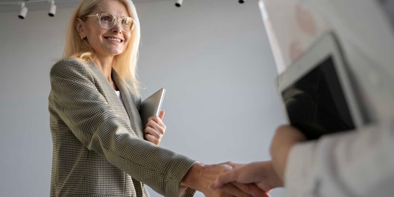 Una mujer rubia sonriente con gafas y un blazer a cuadros le da la mano a otra persona. Sostiene una tableta de plata en su brazo izquierdo. La otra persona, parcialmente visible, también sostiene una tableta. El fondo es claro y neutro, con luces en el techo. La escena transmite un ambiente profesional y agradable.