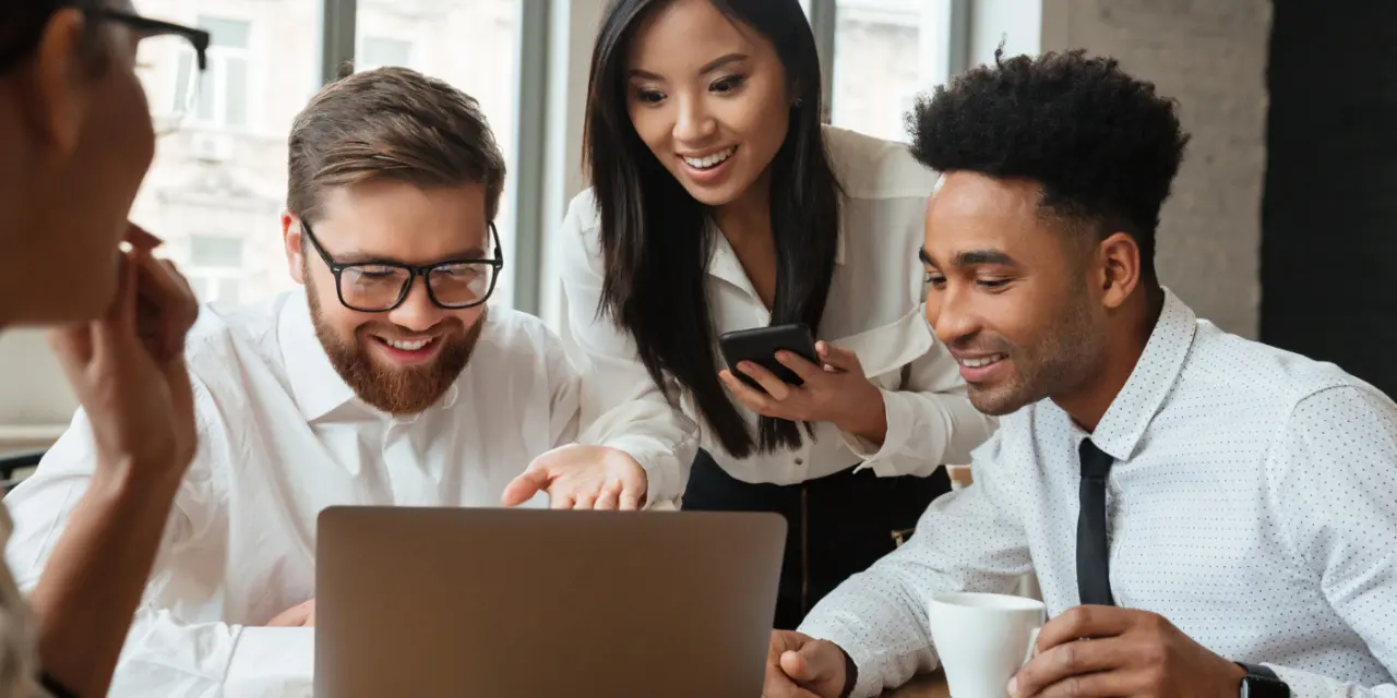 Four people are gathered in a modern workspace, smiling and interacting animatedly in front of a laptop. A bearded man with glasses sits on the left, pointing at the computer screen, while a woman in the center holds a cell phone and watches the screen with interest. On the right, a man in a light shirt and black tie holds a mug and also looks at his laptop. A fourth person is partially visible in the left corner of the image. The space has large windows and natural light, conveying a collaborative and positive atmosphere.