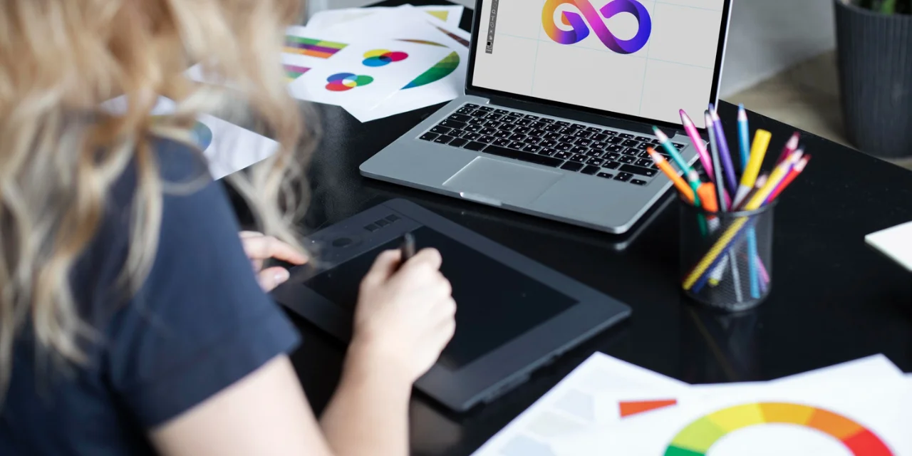 Una persona que usa una tableta gráfica para dibujar o editar un gráfico vectorial, visto parcialmente desde atrás. Delante de él, sobre una mesa negra, hay un portátil con un colorido logotipo en forma de infinito en la pantalla. Sobre la mesa también hay papeles con gráficos de colores, un portalápices con lápices y bolígrafos de colores y otros materiales gráficos. La escena representa un entorno de trabajo creativo, posiblemente relacionado con el diseño gráfico o la identidad visual.